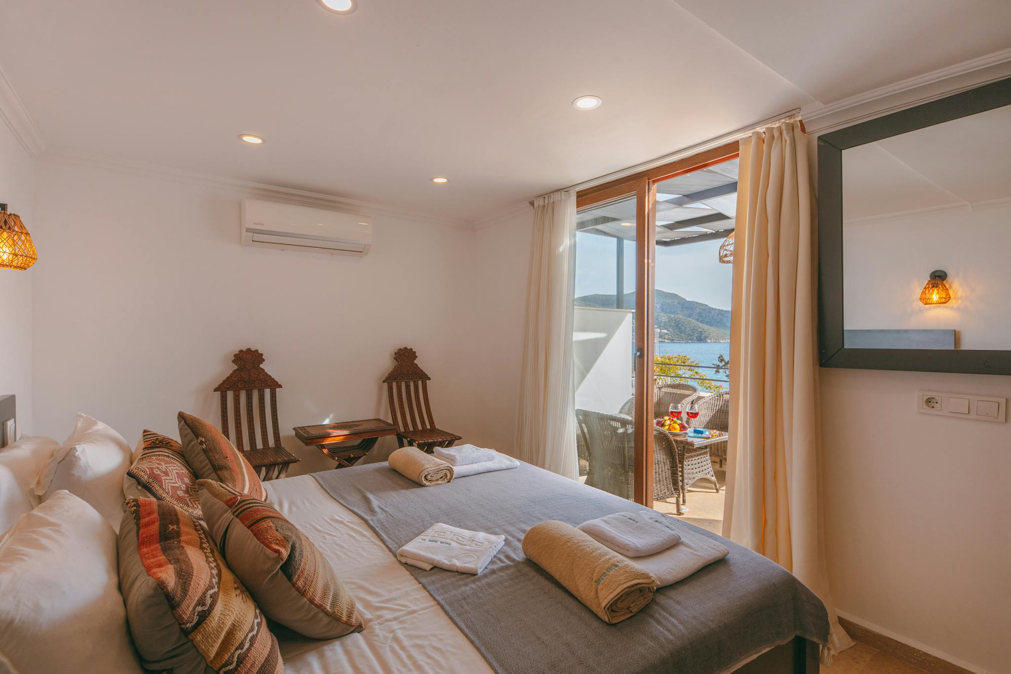 Bright hotel room with bed, chairs, and balcony overlooking the sea.